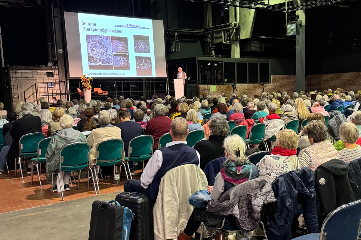 Publikum mit dem Rücken zum Redner. Referent auf der Bühne vor einer Leinwand.